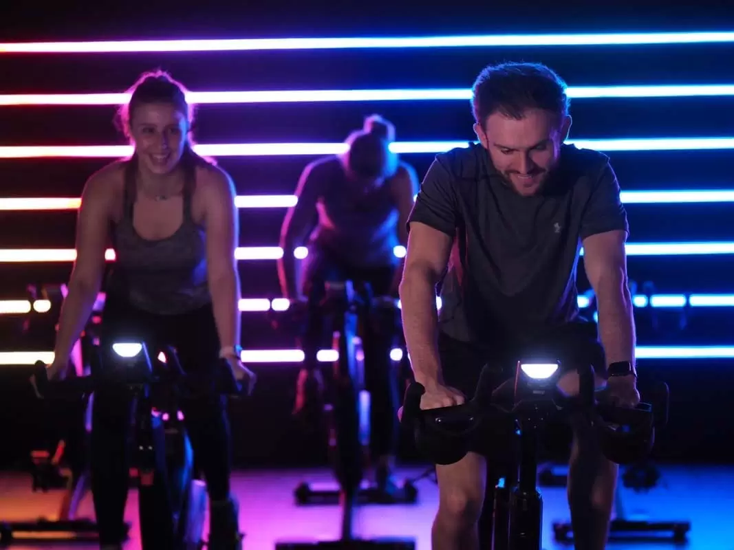 Athletic man and 2 women in their 30s using an indoor bike at The Lensbury's spin studio in Teddington, with use and classes included in their Lensbury membership cost.