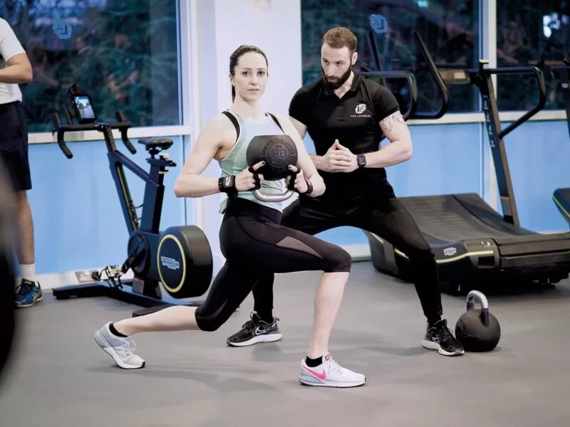 A young woman stretches using a heavy weight whilst a male personal trainer stands alongside within the Lensbury gym in Teddington, where leisure centre classes are available to participate in. The health and fitness centre is open 6:00-22:00 Monday to Thursday, 6:00-21:00 Friday and 7:00-20:00 on weekends.